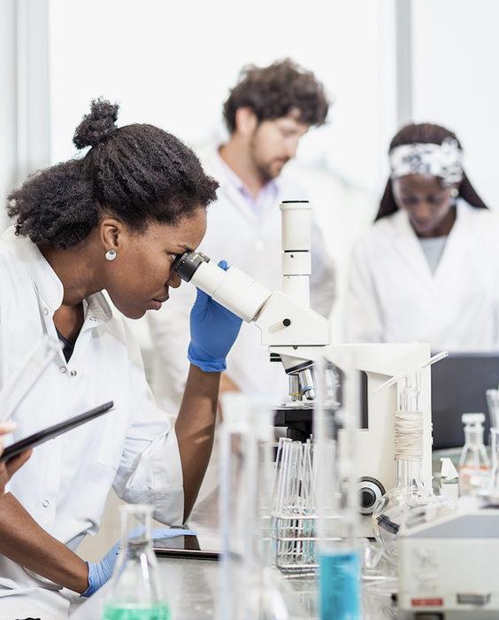 Three people in a lab wearing medical coats, one of them looks through a microscope